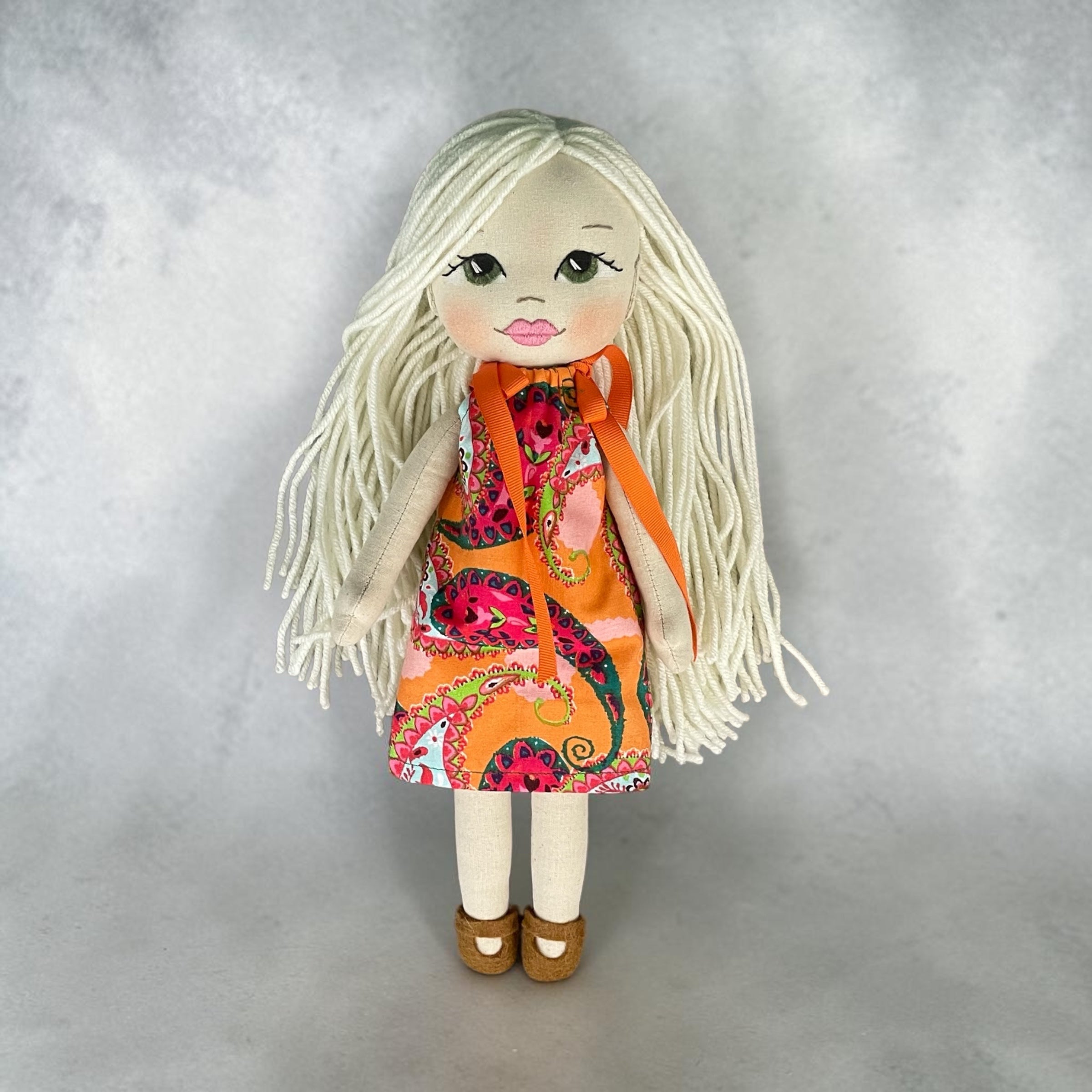 A handmade doll with long white yarn hair, wearing a vibrant orange dress with a multicolor paisley pattern and an orange ribbon, standing against a textured gray background.