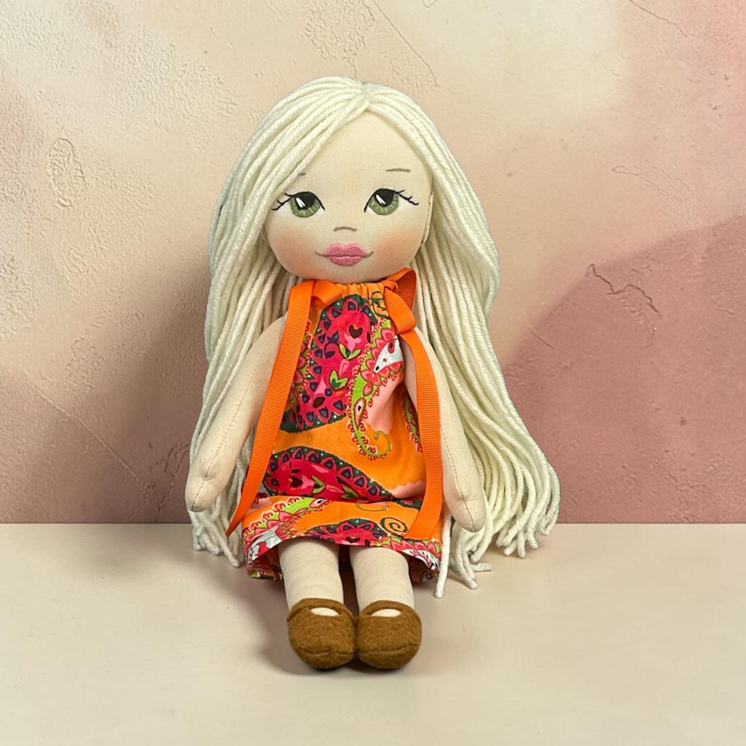 A handmade doll with long white yarn hair, painted green eyes, and a colorful patterned orange dress, sitting on a light beige surface against a textured pinkish-beige wall.