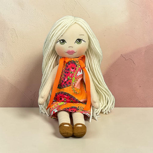 A handmade doll with long white yarn hair, painted green eyes, and a colorful patterned orange dress, sitting on a light beige surface against a textured pinkish-beige wall.